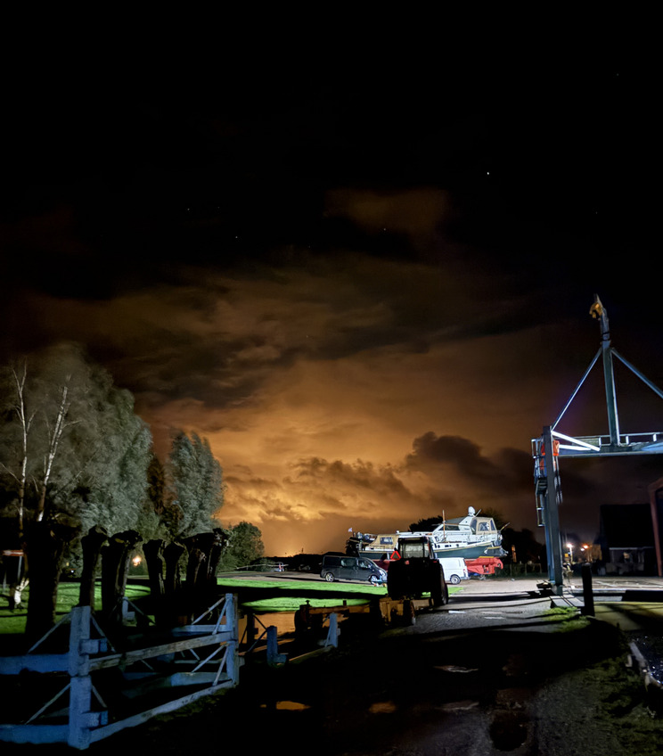 Langelille Nacht Nachtaufnahme eines Hafens mit beleuchteten Wolken am Horizont. Im Vordergrund sind Bäume und eine geschotterte Weg mit einem Traktor und Booten sichtbar. Die Szene vermittelt eine ruhige, maritime Atmosphäre unter dem Nachthimmel.