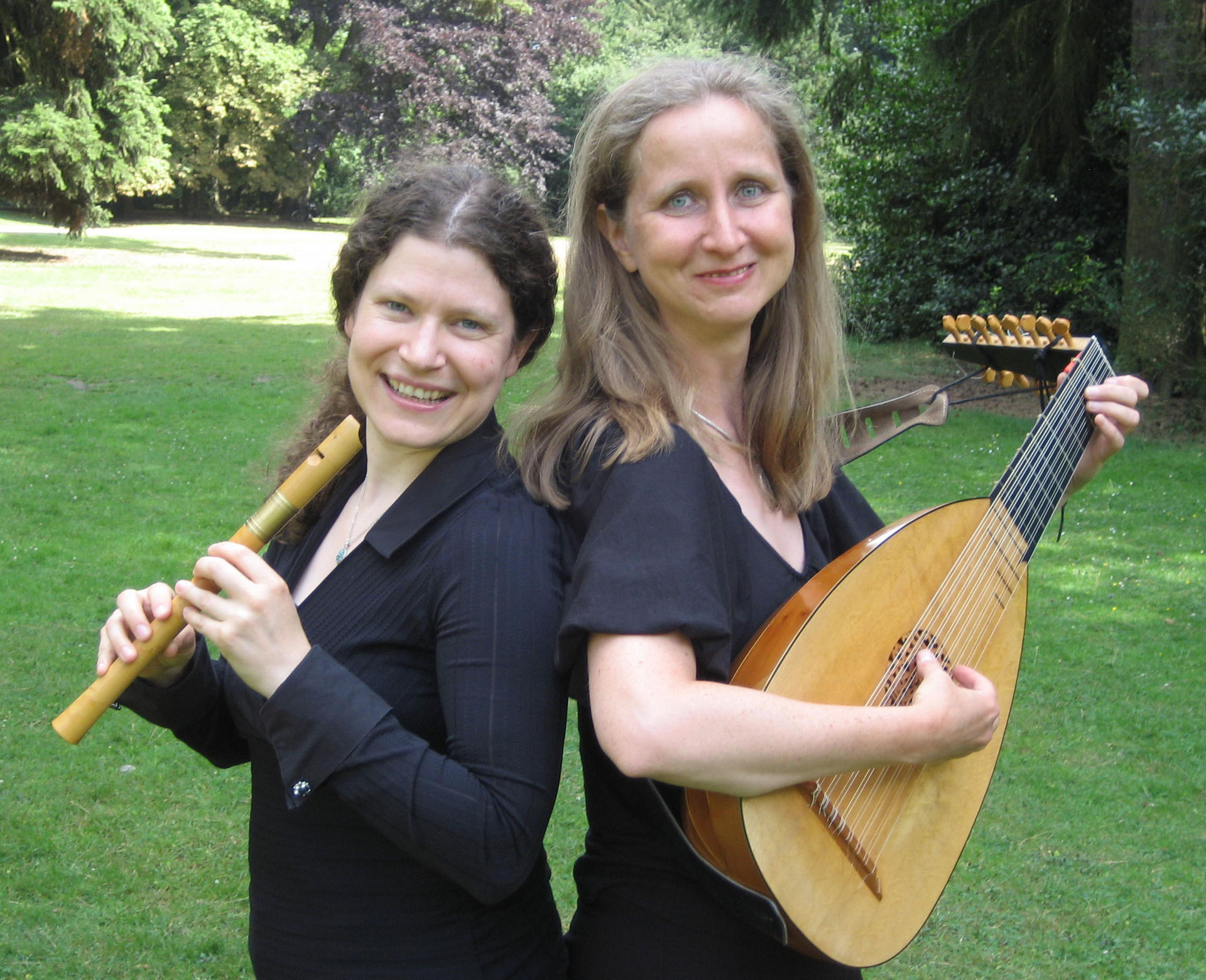 Zwei Musikerinnen stehen im Freien, umgeben von Bäumen. Die erste hält eine Blockflöte, während die zweite eine Laute spielt. Beide tragen schwarze Kleidung und lächeln, was auf eine fröhliche Musikatmosphäre hinweist.
