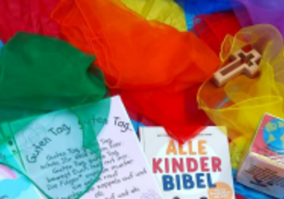 Auf einem bunten Hintergrund liegen verschiedene Tücher in Blau, Rot, Gelb und Grün. Im Vordergrund sind eine Kinderbibel, ein Holzkreuz und Karten mit freundlichen Nachrichten und Zeichnungen zu sehen. Die Gegenstände sind für eine kinderfreundliche Umgebung gestaltet.