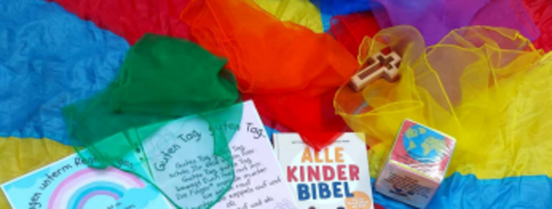 Auf einem bunten Hintergrund liegen verschiedene Tücher in Blau, Rot, Gelb und Grün. Im Vordergrund sind eine Kinderbibel, ein Holzkreuz und Karten mit freundlichen Nachrichten und Zeichnungen zu sehen. Die Gegenstände sind für eine kinderfreundliche Umgebung gestaltet.