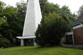 Ein moderner, schmaler Turm mit vertikalen weißen Lamellen steht auf einem grünen Rasen. Umgeben von Bäumen und niedrigem Strauch, ist der Turm Teil eines architektonischen Ensembles, das in einem natürlichen Umfeld integriert ist.