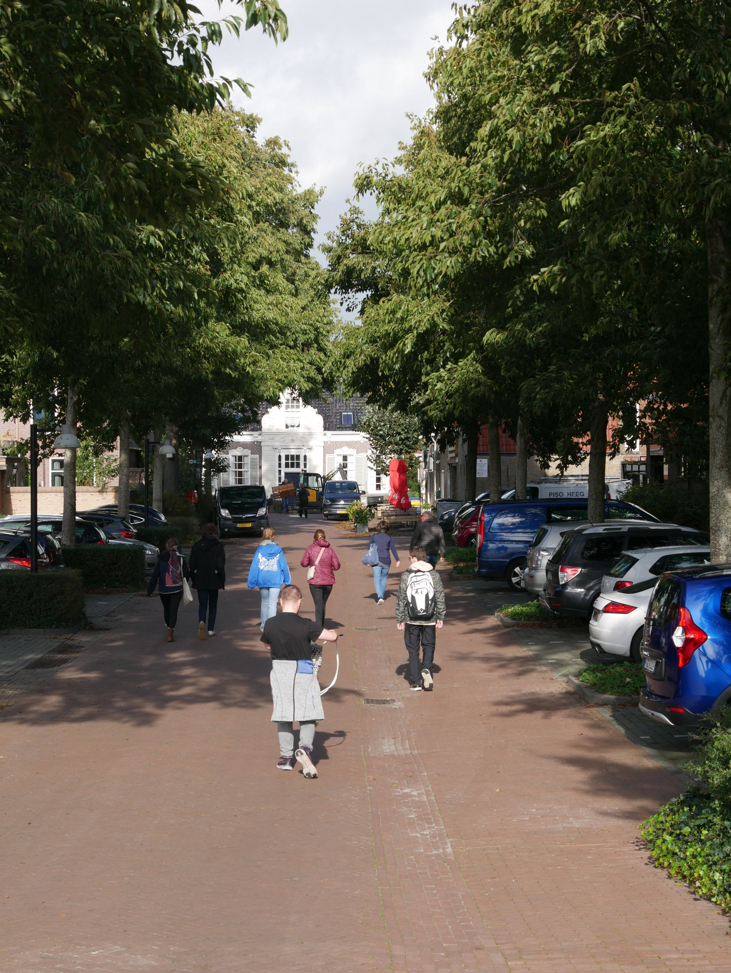 Eine Gruppe Menschen geht eine grade Straße hinunter die Richtung Stadt führt. Die Sonne scheint.