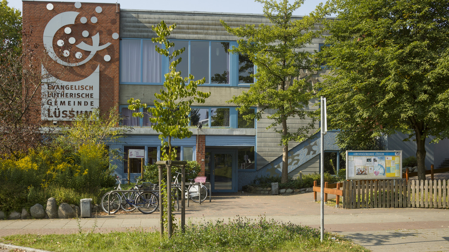 Das Bild zeigt das Gebäude der evangelisch-lutherischen Gemeinde in Lüsum. Die Fassade ist in neutralen Farben gestaltet, mit großen Fenstern. Vor dem Gebäude stehen einige Fahrräder und Bäume, die das Umfeld begrünen. An einer Wand ist ein Gestaltungselement sichtbar.