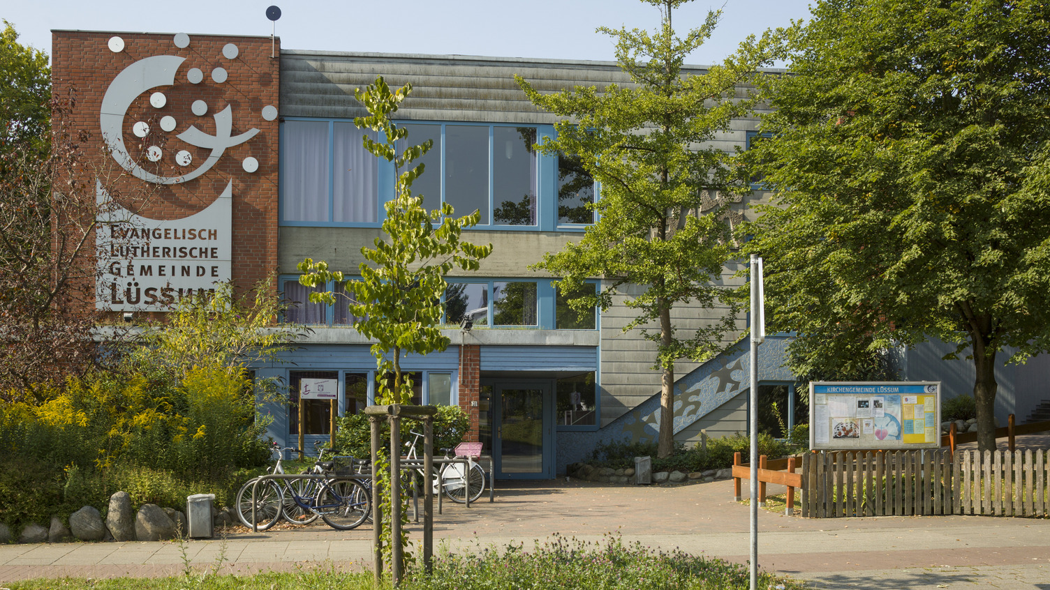 Das Bild zeigt das Gebäude der evangelisch-lutherischen Gemeinde in Lüsum. Die Fassade ist in neutralen Farben gestaltet, mit großen Fenstern. Vor dem Gebäude stehen einige Fahrräder und Bäume, die das Umfeld begrünen. An einer Wand ist ein Gestaltungselement sichtbar.