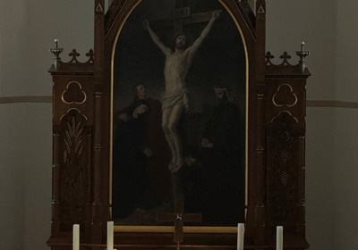 Ein Kirchenaltar mit einem großen Gemälde der Kreuzigung Jesu im Hintergrund. Vor dem Bild stehen drei Kerzen auf einem Holzaltar. Ein aufgeschlagenes Buch ist beleuchtet, was eine ruhige und feierliche Atmosphäre schafft. Hinter dem Altar sind farbige Fenster zu erkennen.