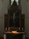 Ein Kirchenaltar mit einem großen Gemälde der Kreuzigung Jesu im Hintergrund. Vor dem Bild stehen drei Kerzen auf einem Holzaltar. Ein aufgeschlagenes Buch ist beleuchtet, was eine ruhige und feierliche Atmosphäre schafft. Hinter dem Altar sind farbige Fenster zu erkennen.
