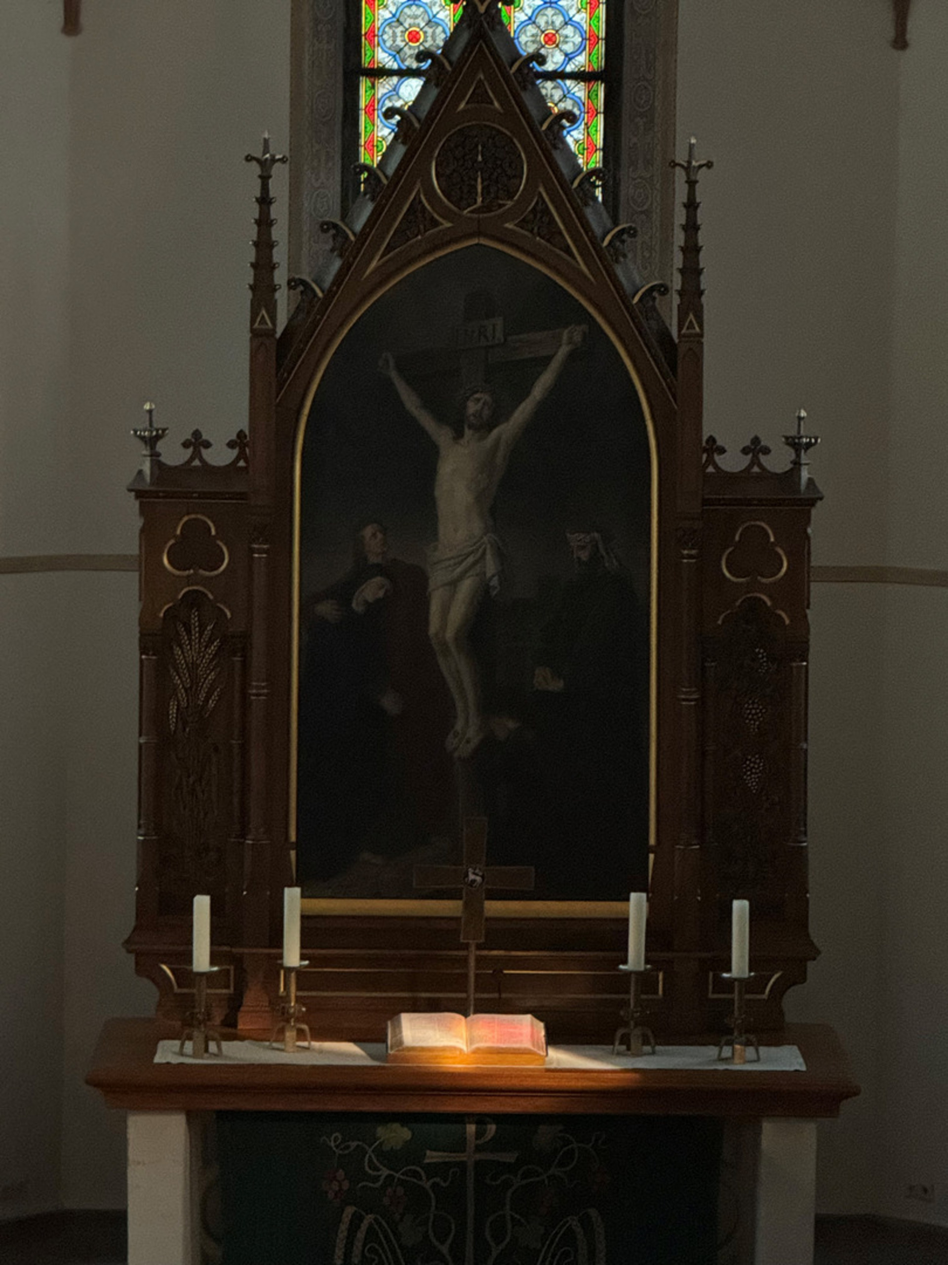 Ein Kirchenaltar mit einem großen Gemälde der Kreuzigung Jesu im Hintergrund. Vor dem Bild stehen drei Kerzen auf einem Holzaltar. Ein aufgeschlagenes Buch ist beleuchtet, was eine ruhige und feierliche Atmosphäre schafft. Hinter dem Altar sind farbige Fenster zu erkennen.