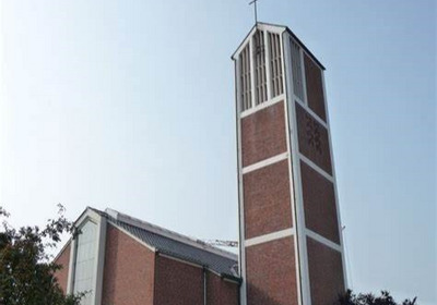 Eine moderne Kirche ist in der Bildmitte zu sehen. Ihr schlanker, vierseitiger Turm hat oben ein Kreuz. Das Gebäude ist rot gemauert und von Bäumen umgeben. Der Himmel ist klar und blau.