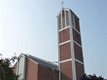 Eine moderne Kirche ist in der Bildmitte zu sehen. Ihr schlanker, vierseitiger Turm hat oben ein Kreuz. Das Gebäude ist rot gemauert und von Bäumen umgeben. Der Himmel ist klar und blau.