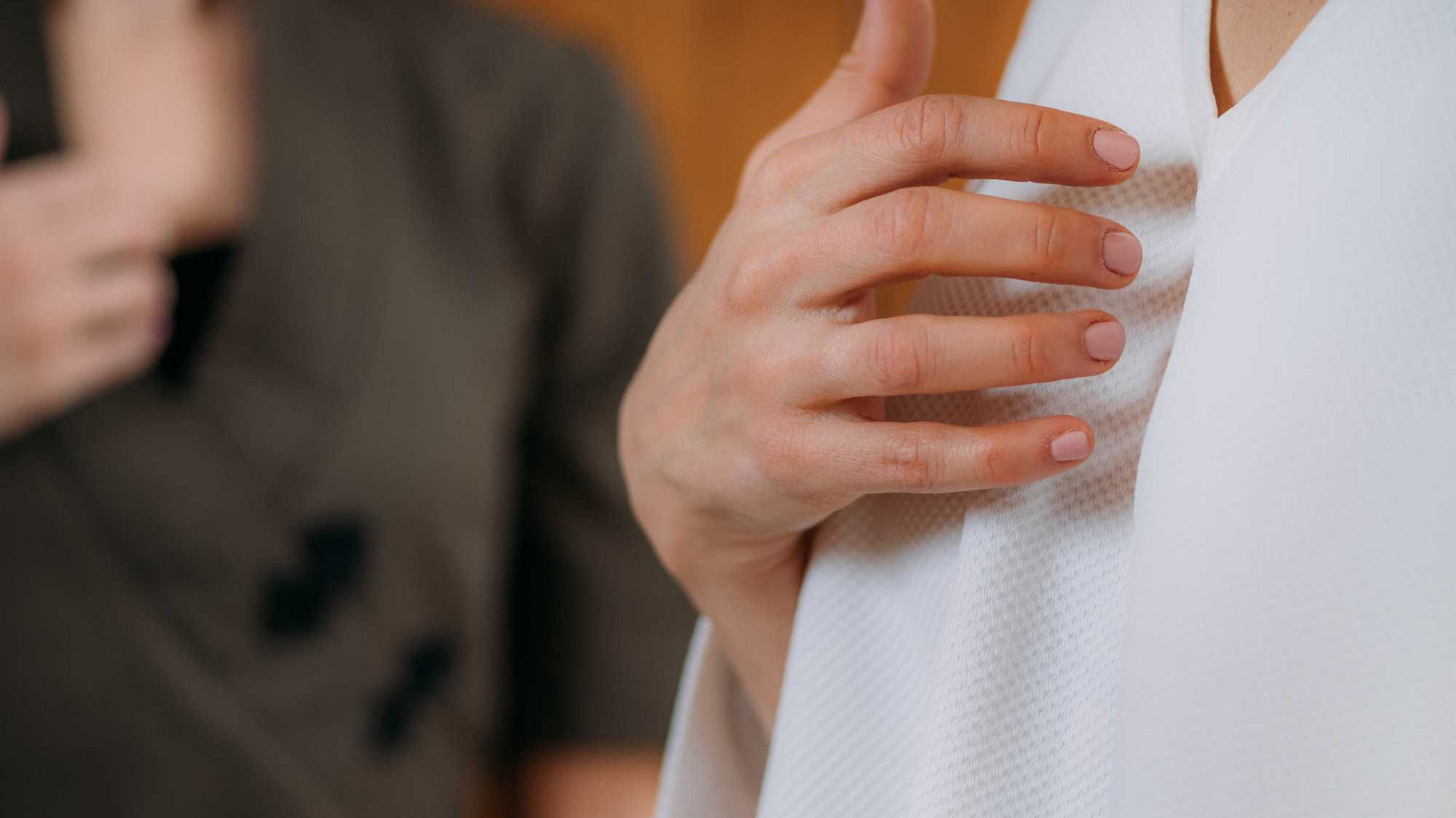 Zwei Personen stehen im Bild. Eine Frau trägt ein weißes Oberteil und legt ihre Hand auf die Brust. Die andere Person im Hintergrund trägt ein dunkles Oberteil und hat ebenfalls eine Hand auf der Brust. Die Atmosphäre wirkt ruhig und förderlich für eine harmonische Interaktion.