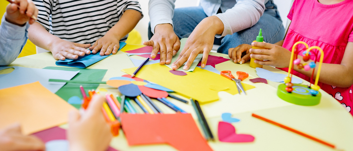 Kinder und Erzieherin sitzen an einem Tisch und basteln. Es sind keine Gesichter zu sehen.