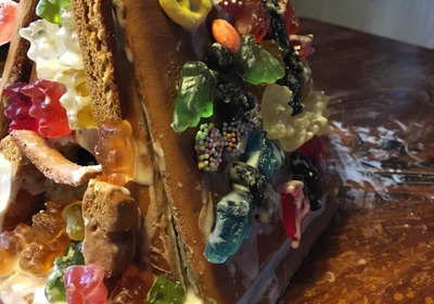 Ein bunt dekoriertes Lebkuchenhaus, überzogen mit verschiedenen Gummibärchen, Süßigkeiten und bunter Glasur. Die Süßigkeiten sind vielfältig angeordnet und verleihen dem Haus ein fröhliches, festliches Aussehen. Der Hintergrund ist aus Holz, was dem Bild eine gemütliche Stimmung verleiht.