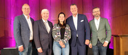 Fünf Personen stehen auf einer Bühne vor einer hölzernen Wand mit lila Beleuchtung. Sie lächeln und sind formell gekleidet. Die mittlere Person trägt einen bunten Pullover und hält die Hände vor sich. Die anderen Personen sind in Anzügen gekleidet.