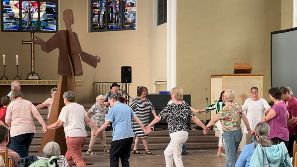 Eine Gruppe von Menschen unterschiedlicher Altersgruppen tanzt in einem Kirchenraum im Kreis. In der Mitte steht eine große Holzskulptur. Die Atmosphäre ist freundlich und lebhaft, die Teilnehmer halten sich an den Händen und lächeln. Bunte Kirchenfenster sind im Hintergrund zu sehen.
