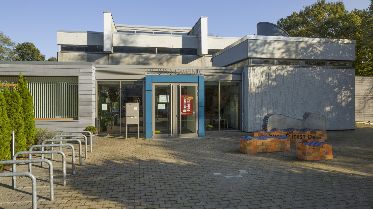 Das Bild zeigt das moderne Gebäude eines Gemeindezentrums mit einer blauen Eingangstür. Vor dem Gebäude befindet sich ein gepflasterter Platz mit Fahrradbügeln und einem bunten Sitzbereich aus Steinen. Die Umgebung ist hell und sonnig, mit Bäumen im Hintergrund.