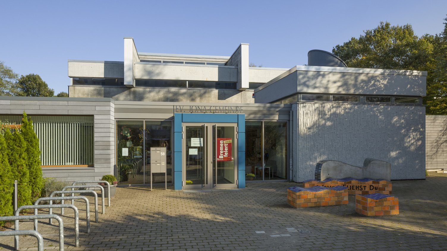 Das Bild zeigt das moderne Gebäude eines Gemeindezentrums mit einer blauen Eingangstür. Vor dem Gebäude befindet sich ein gepflasterter Platz mit Fahrradbügeln und einem bunten Sitzbereich aus Steinen. Die Umgebung ist hell und sonnig, mit Bäumen im Hintergrund.