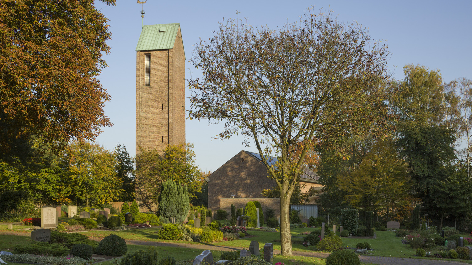 Ein Blick auf einen Friedhof mit sorgfältig gepflegten Gräbern und bunten Blumenbeeten. Im Hintergrund steht ein hoher, rechteckiger Kirchturm mit grünem Dach und einer Wetterfahne. Umgeben von Bäumen.