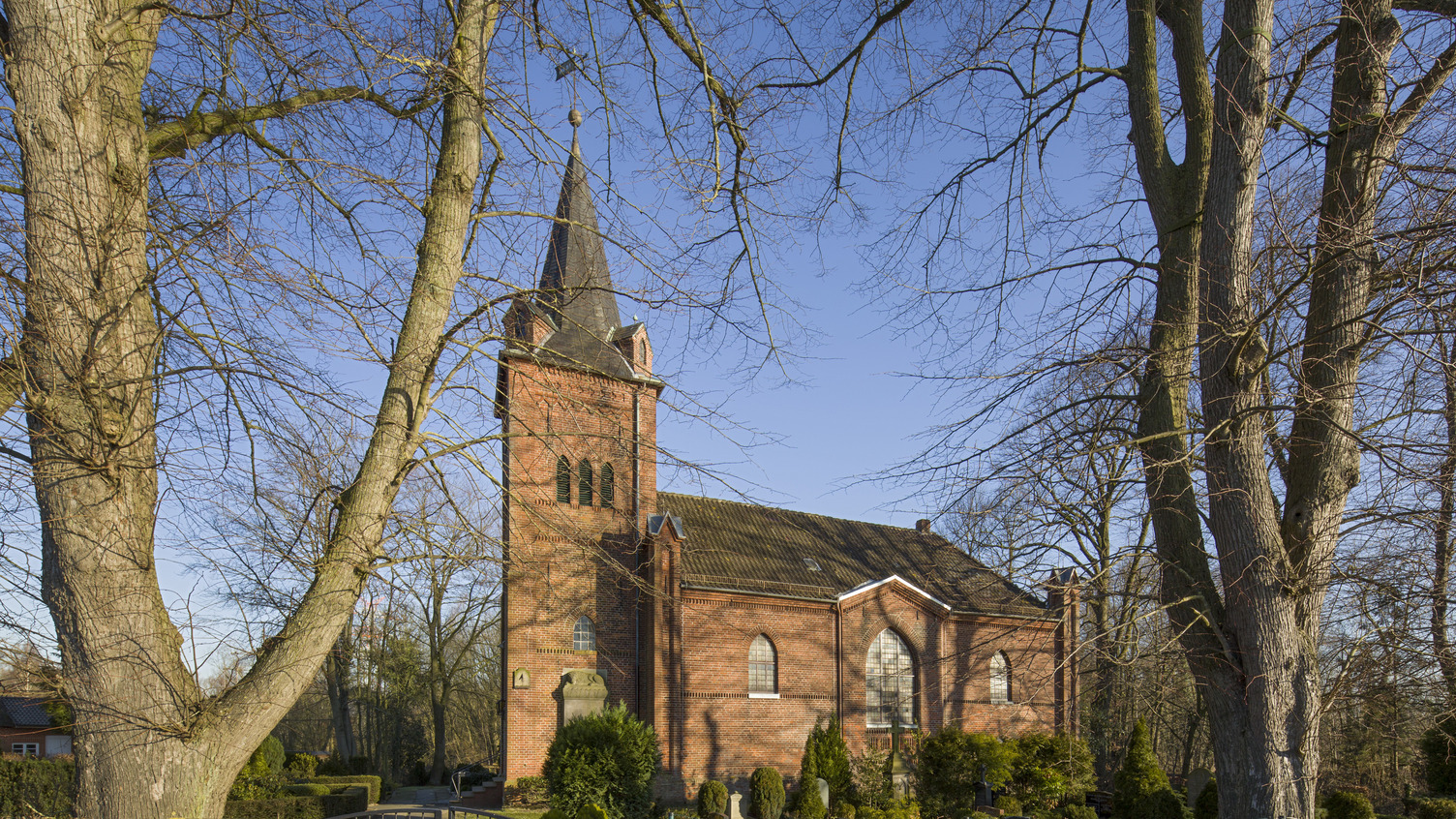 Eine sich im Sonnenlicht präsentierende Backsteinkirche mit einem hohen, spitz zulaufenden Turm. Der Kirchenbau ist von Bäumen und einem gepflegten Garten umgeben, der durch ein Tor zugänglich ist.