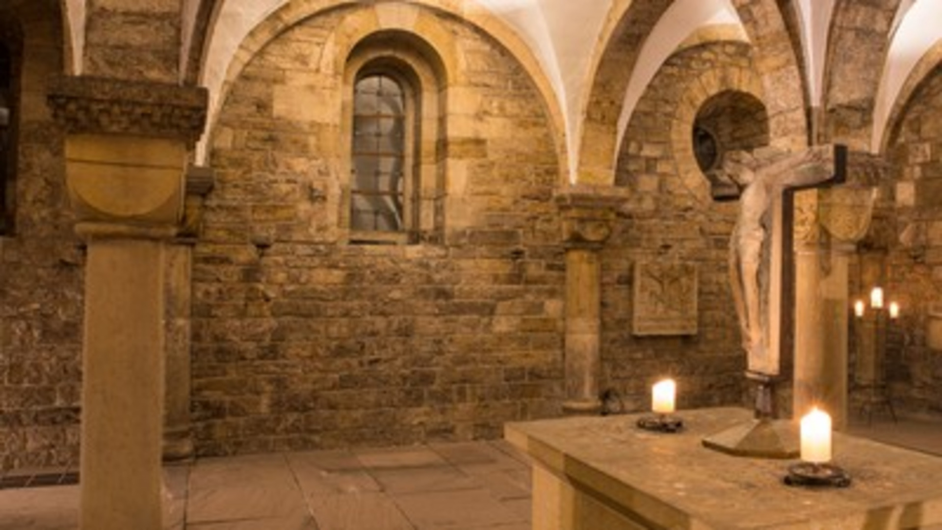 Im Bild ist ein historischer, steinerner Raum mit gewölbten Decken zu sehen. In der Mitte steht ein Tisch mit Kerzen, und an einer Säule hängt eine Statue von Christus am Kreuz. Die Wände sind aus unbehandeltem Stein, was eine mystische Atmosphäre schafft.