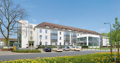 Das Bild zeigt das Gebäude der Roland-Klinik, ein modernes, mehrstöckiges Krankenhaus. Im Vordergrund blühen gelbe Narzissen, und auf dem Parkplatz stehen mehrere Autos. Der Himmel ist blau mit wenigen Wolken.