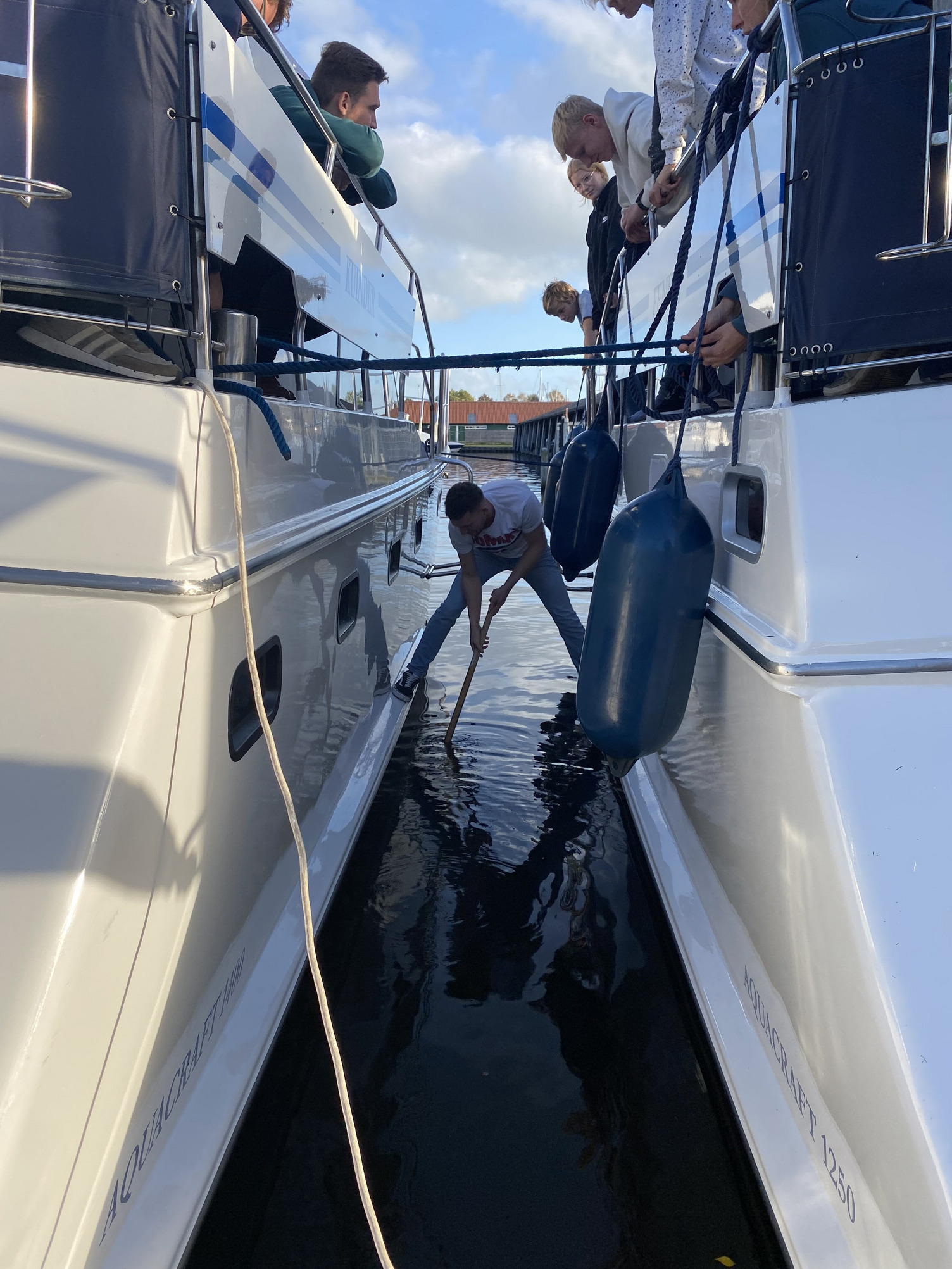 Eine Person balanciert zwischen zwei Booten und versucht eine über Bord gegangene Brille aus dem Hafen zu fischen.