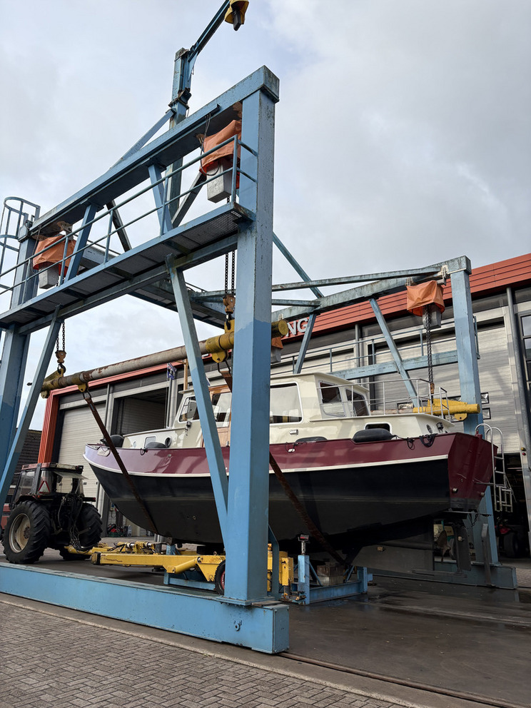 Fliegendes Boot Ein Boot wird mit einem Kran auf einem Schiffslift angehoben. Der Kran ist Teil einer großen blauen Struktur, die im Freien steht. Im Hintergrund sind verschiedene Gebäude sowie ein Traktor zu sehen. Die Umgebung wirkt industriell und funktional.
