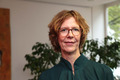 Eine Person mit lockigem, blondem Haar und Brille steht in einem hellen Raum mit Pflanzen im Hintergrund. Sie trägt ein dunkles Oberteil und blickt direkt in die Kamera.