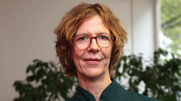 Eine Person mit lockigem, blondem Haar und Brille steht in einem hellen Raum mit Pflanzen im Hintergrund. Sie trägt ein dunkles Oberteil und blickt direkt in die Kamera.