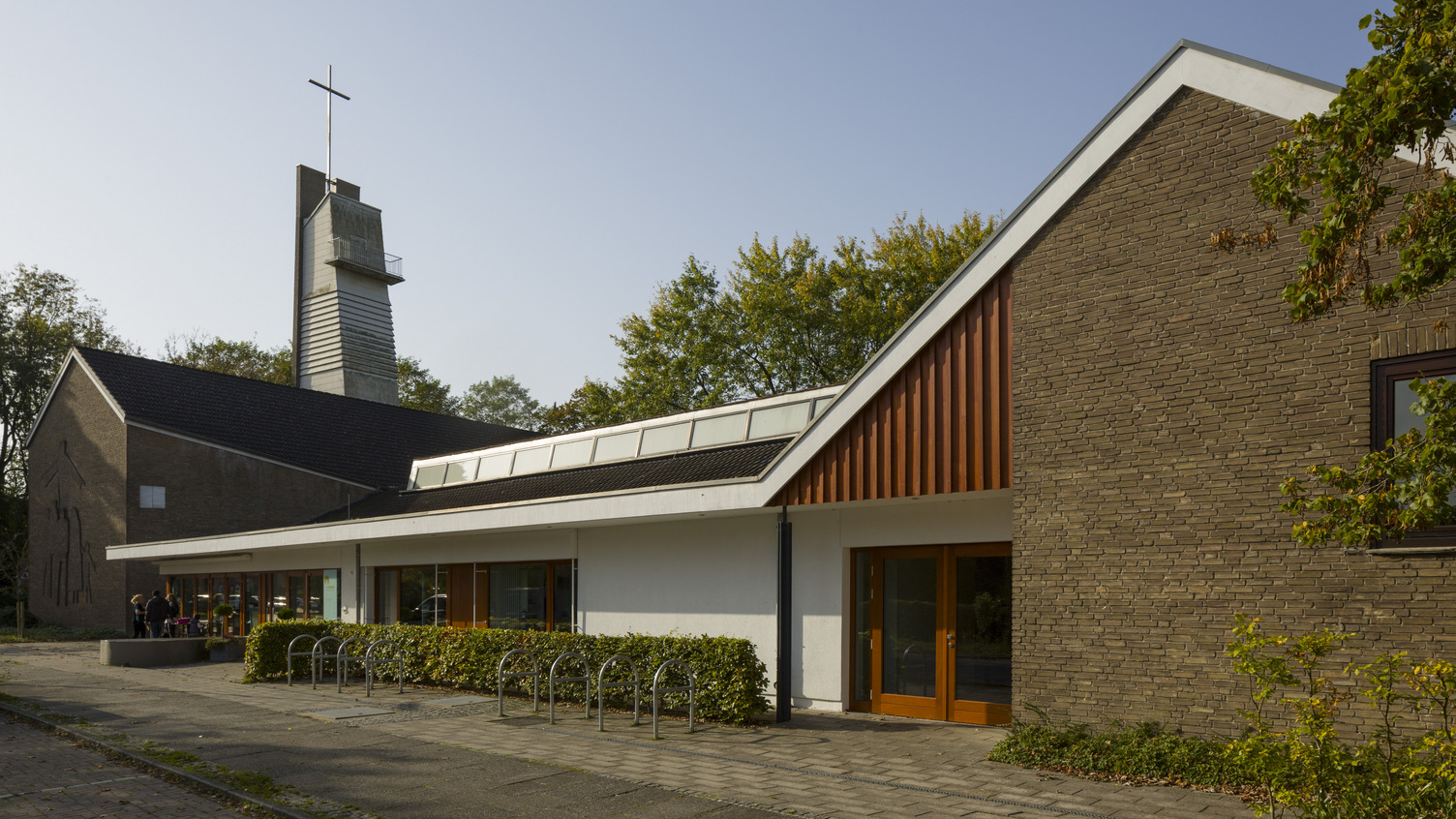 Eine moderne Kirche mit einem hohen Glockenturm und einem Kreuz an der Spitze. Die Fassade ist eine Kombination aus Ziegel und weißen Wänden. Es gibt Pflanzen entlang des Gehwegs. Der Himmel ist klar und sonnig.