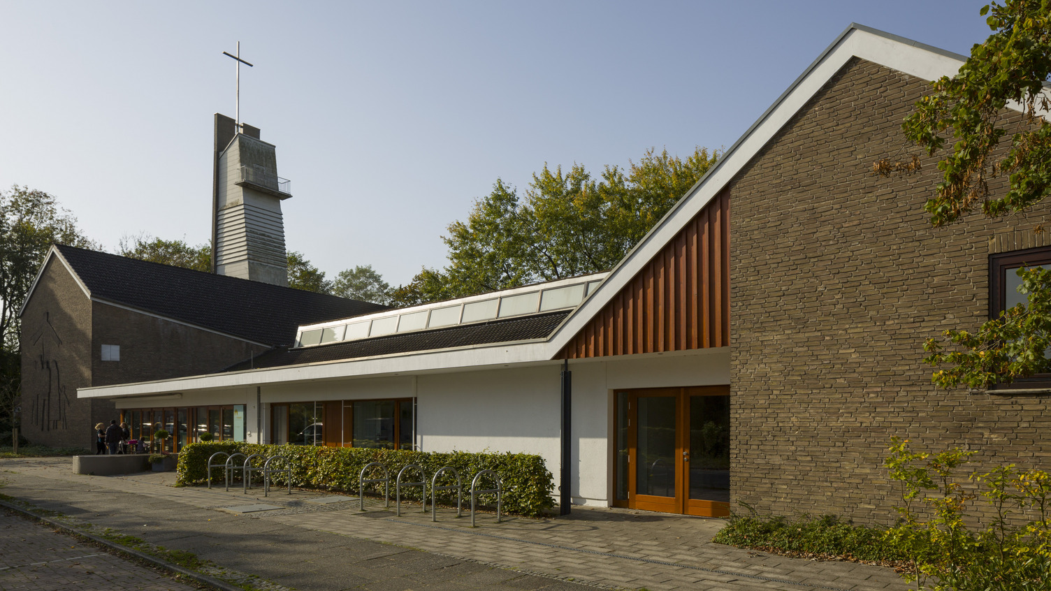 Eine moderne Kirche mit einem hohen Glockenturm und einem Kreuz an der Spitze. Die Fassade ist eine Kombination aus Ziegel und weißen Wänden. Es gibt Pflanzen entlang des Gehwegs. Der Himmel ist klar und sonnig.