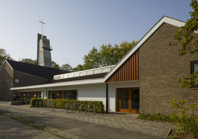 Eine moderne Kirche mit einem hohen Glockenturm und einem Kreuz an der Spitze. Die Fassade ist eine Kombination aus Ziegel und weißen Wänden. Es gibt Pflanzen entlang des Gehwegs. Der Himmel ist klar und sonnig.