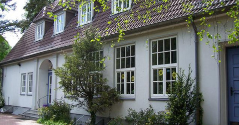 Ein zweigeschossiges, helles Gebäude mit einem Satteldach. Große Fenster lassen viel Licht einfallen. Vor dem Gebäude befinden sich Sträucher und Bäume. Der Eingangsbereich ist durch eine kleine Treppe zugänglich. Die Umgebung wirkt grün und einladend.