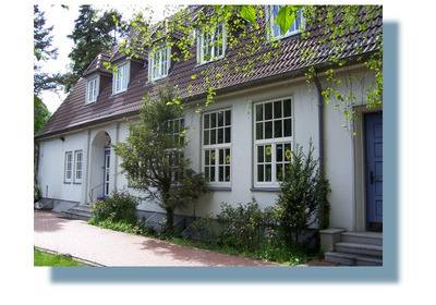 Ein zweigeschossiges, helles Gebäude mit einem Satteldach. Große Fenster lassen viel Licht einfallen. Vor dem Gebäude befinden sich Sträucher und Bäume. Der Eingangsbereich ist durch eine kleine Treppe zugänglich. Die Umgebung wirkt grün und einladend.