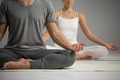 Zwei Personen sitzen im Schneidersitz auf einer Matte und praktizieren Yoga. Der Mann trägt ein graues T-Shirt und Hosen, während die Frau ein weißes Oberteil und helle Leggings trägt. Beide sind entspannt und in meditativer Haltung.