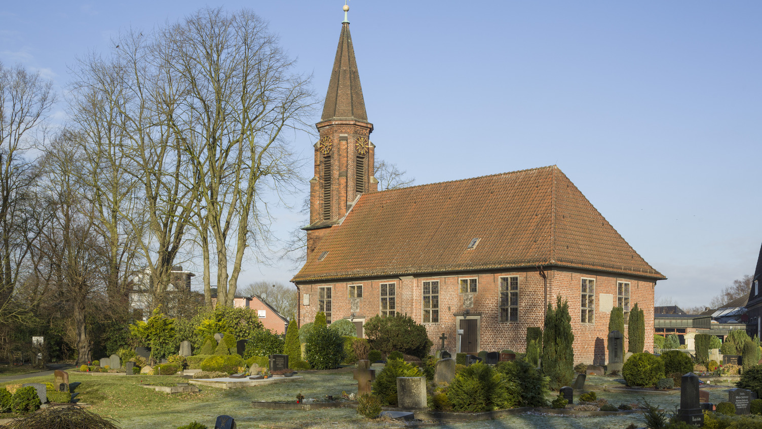 Ein Ziegeldach sitzt auf einer Kirche mit einem schlanken Turm, umgeben von einem Friedhof. Die Umgebung ist grün mit mehreren Bäumen und gut gepflegten Gräbern. Ein klarer blauer Himmel bildet den Hintergrund.