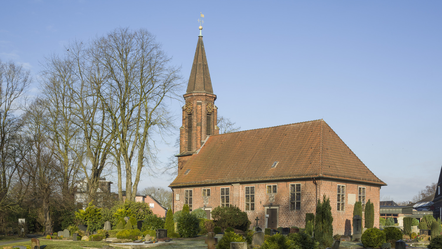 Ein Ziegeldach sitzt auf einer Kirche mit einem schlanken Turm, umgeben von einem Friedhof. Die Umgebung ist grün mit mehreren Bäumen und gut gepflegten Gräbern. Ein klarer blauer Himmel bildet den Hintergrund.