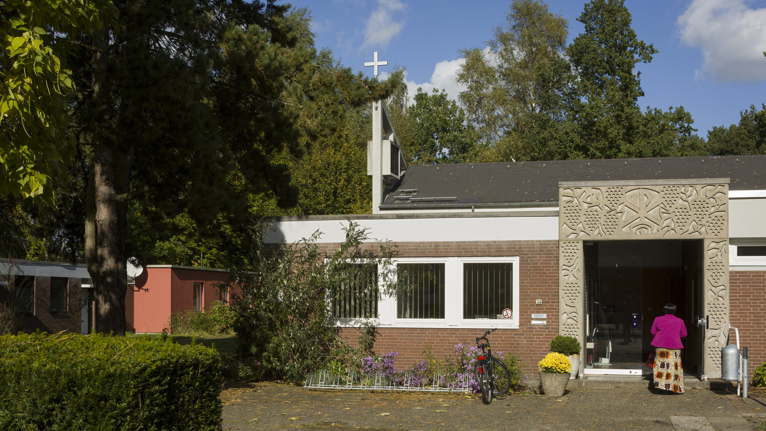 Ein modernes Gebäude mit einer kunstvoll verzierten Fassade und einem Kreuz auf dem Dach. Im Vordergrund steht eine Person in pinkem Oberteil, die zur Eingangstür schreitet. Bunte Blumen und ein Fahrrad sind ebenfalls sichtbar. Die Umgebung ist von Bäumen umgeben.