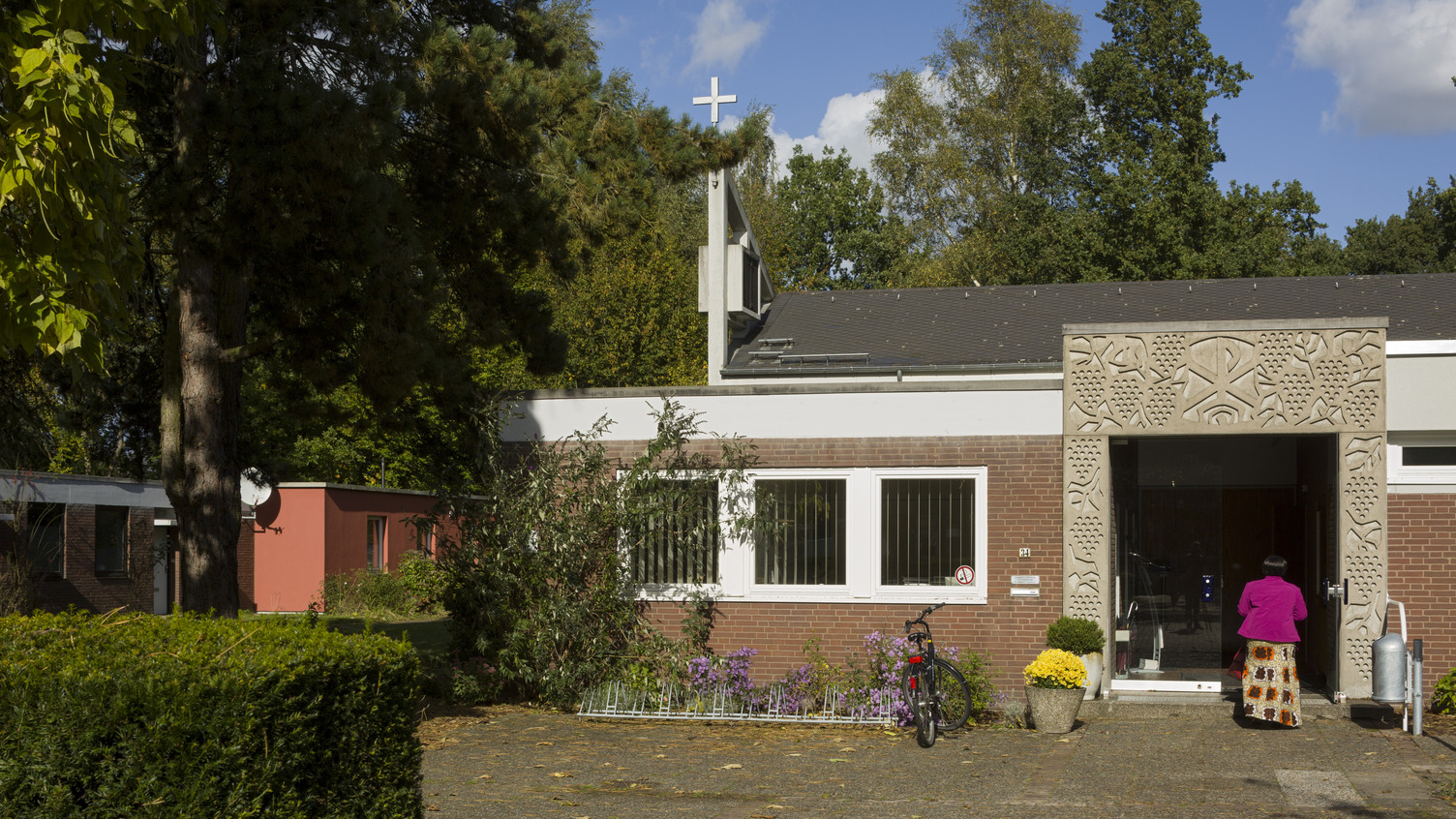 Ein modernes Gebäude mit einer kunstvoll verzierten Fassade und einem Kreuz auf dem Dach. Im Vordergrund steht eine Person in pinkem Oberteil, die zur Eingangstür schreitet. Bunte Blumen und ein Fahrrad sind ebenfalls sichtbar. Die Umgebung ist von Bäumen umgeben.