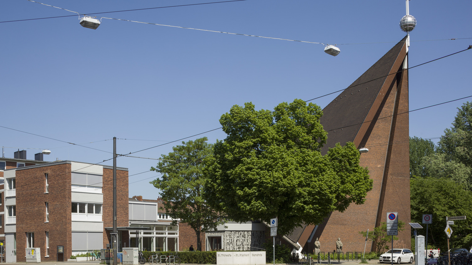 Ein modernes, schräg gebautes Kirchgebäude mit einem Kreuz und einer Kugel auf dem Dach. Vor der Kirche steht ein großer Baum, und daneben befinden sich ein paar Menschen und Fahrräder. Die Umgebung zeigt auch einige Gebäude und einen klaren blauen Himmel.