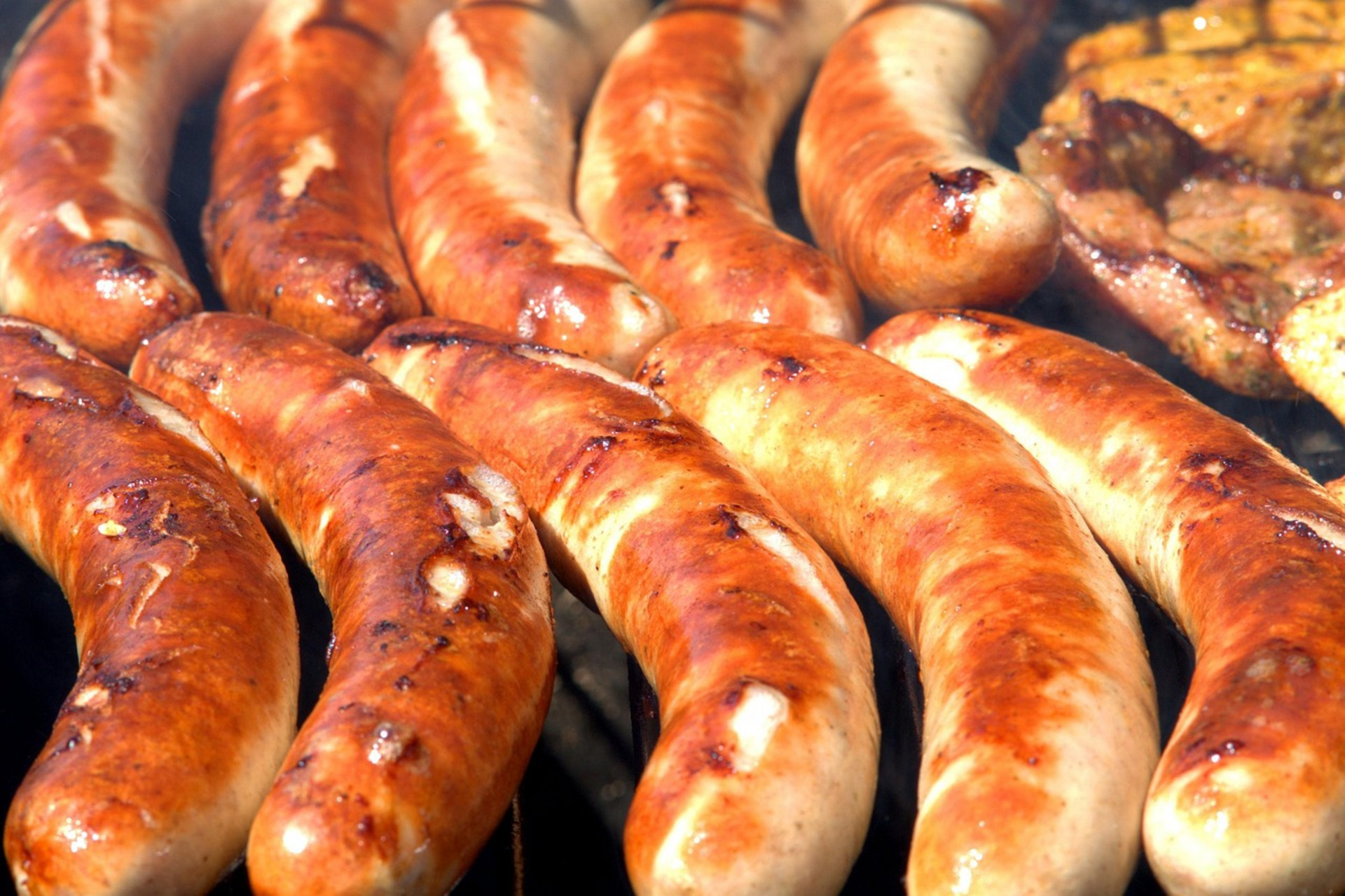 Gegrillte Würstchen liegen auf einem Grill. Sie sind goldbraun und leicht knusprig, mit rauchigem Aroma. Im Hintergrund sind weitere Grillgerichte zu sehen.