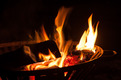 Ein loderndes Feuer im Grill mit glühenden Holzscheiten. Die Flammen tanzen in der Dunkelheit, während die roten Glutstücke sichtbar sind.