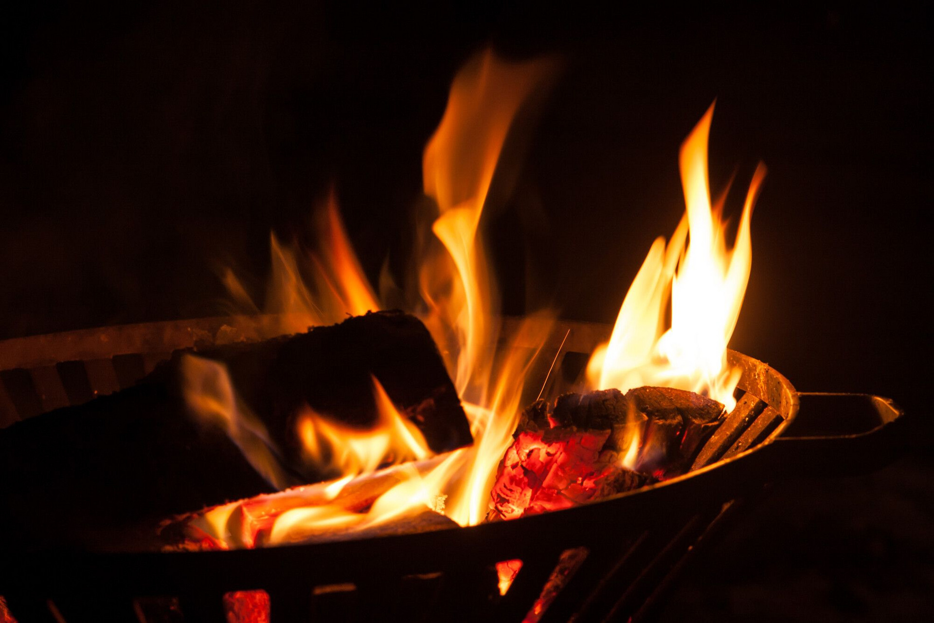 Ein loderndes Feuer im Grill mit glühenden Holzscheiten. Die Flammen tanzen in der Dunkelheit, während die roten Glutstücke sichtbar sind.