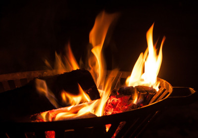 Ein loderndes Feuer im Grill mit glühenden Holzscheiten. Die Flammen tanzen in der Dunkelheit, während die roten Glutstücke sichtbar sind.