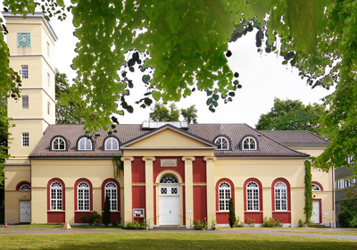 Das Bild zeigt ein historisches Gebäude mit einer auffälligen Fassade in Gelb und Rot. Vor dem Haupteingang befinden sich Säulen und große Fenster. Links ist ein Uhrenturm zu sehen, umgeben von Bäumen. Die Umgebung ist grün und einladend.