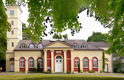 Das Bild zeigt ein historisches Gebäude mit einer auffälligen Fassade in Gelb und Rot. Vor dem Haupteingang befinden sich Säulen und große Fenster. Links ist ein Uhrenturm zu sehen, umgeben von Bäumen. Die Umgebung ist grün und einladend.