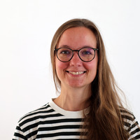 Eine Frau mit langen, braunen Haaren lächelt in die Kamera. Sie trägt eine schwarz-weiß gestreifte Bluse und Brille. Der Hintergrund ist einfarbig weiß.