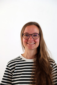 Eine Frau mit langen, braunen Haaren lächelt in die Kamera. Sie trägt eine schwarz-weiß gestreifte Bluse und Brille. Der Hintergrund ist einfarbig weiß.