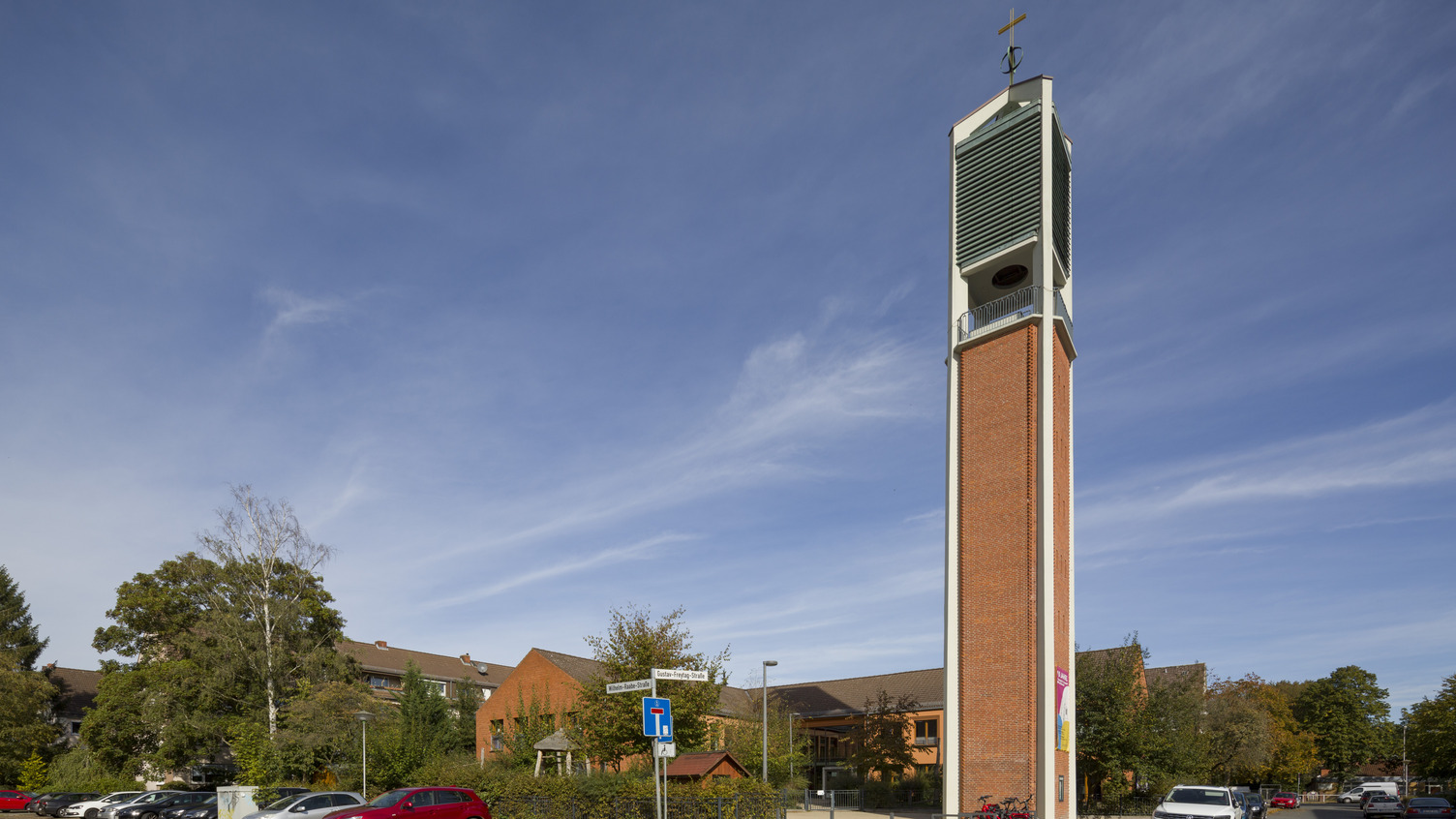 Ein hoher, schlanker Kirchturm aus roten Ziegeln mit einem Kreuz an der Spitze ist sichtbar. Er steht in der Nähe eines Gebäudes, das von Bäumen umgeben ist. Im Vordergrund sind mehrere parkende Autos zu sehen. Der Himmel ist blau mit einigen Wolken.