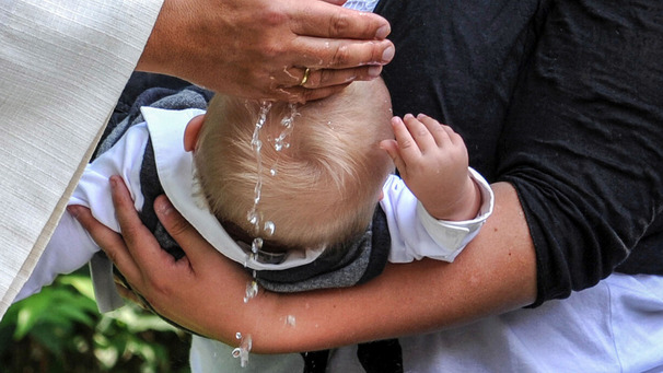 Das Foto zeigt ein kleines Kind auf dem Arm seiner Mutter, das gerade getauft wird.