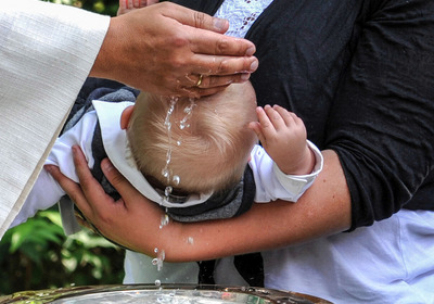 Das Foto zeigt ein kleines Kind auf dem Arm seiner Mutter, das gerade getauft wird.